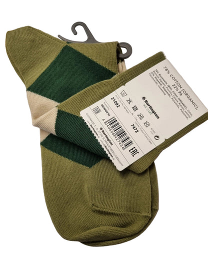 A pair of knit socks with a green and beige argyle pattern, shown with a white paper tag attached. The socks are photographed on a white sheet of paper with a wooden surface visible in the top-left corner.