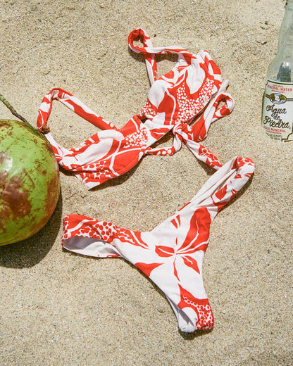 Off-model flat lay of the LSPACE Hibiscus Bloom Rocky Bikini Bottom and Mako Bikini Top arranged on sandy beach beside a coconut.