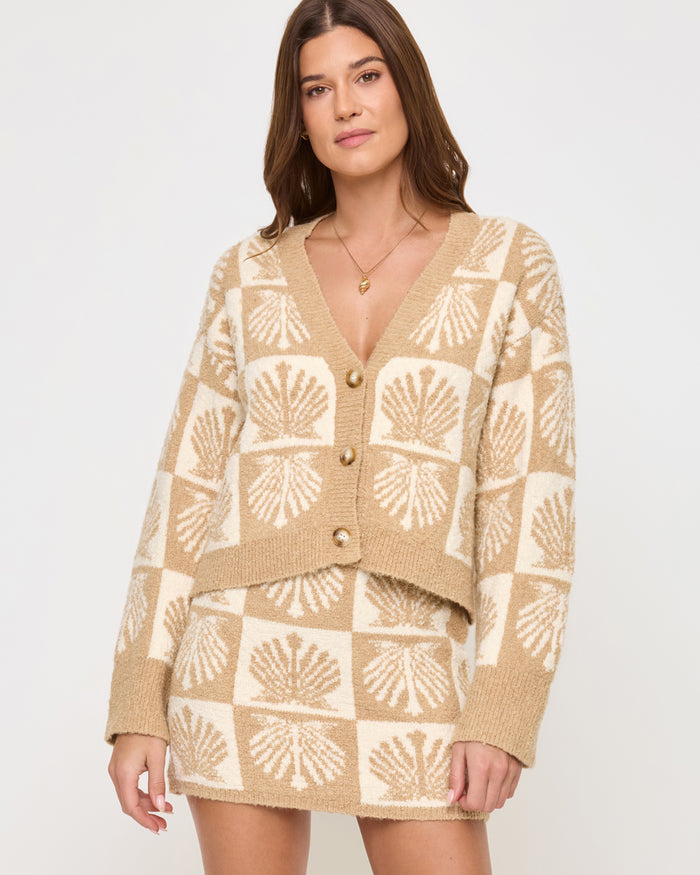 Close front view of a model wearing the LSPACE Shell Be There Skirt in seashell jacquard, showing the knit mini skirt with a checkered seashell pattern in tan and cream, paired with the coordinating cardigan.