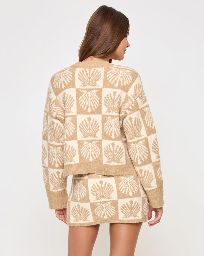 Back view of a model wearing the LSPACE Shell Be There Skirt in seashell jacquard, showing the knit mini skirt with the checkered seashell pattern in tan and cream, styled with the matching cardigan.